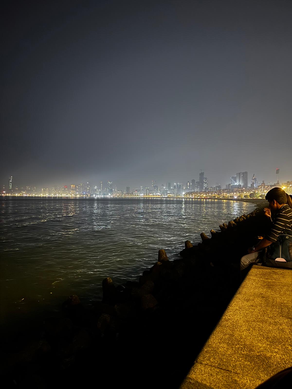 People sitting beside the sea at Marine Drive with Mumbai’s waterfront lights at night
