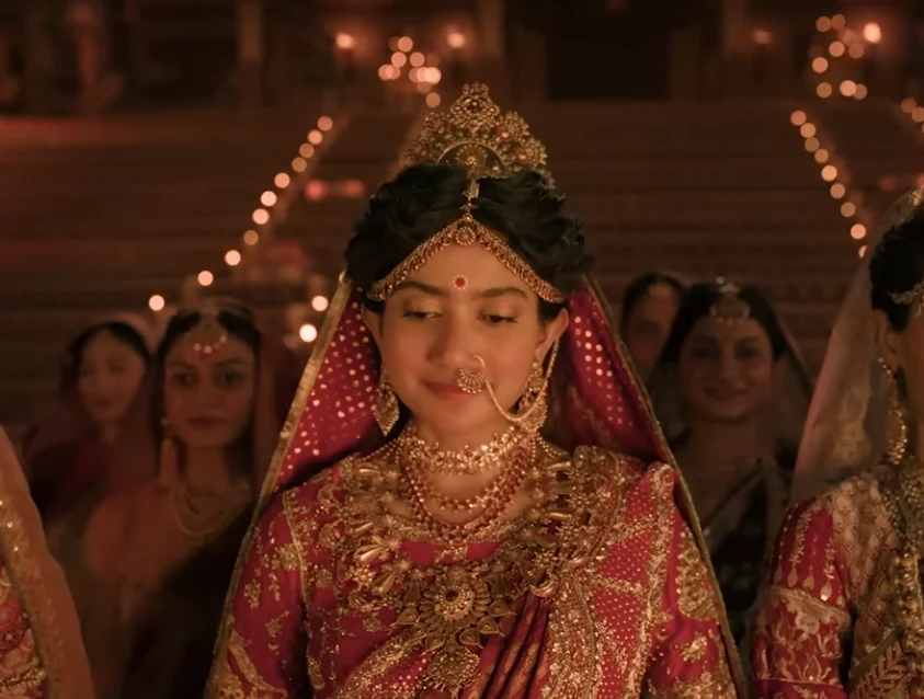 A woman dressed as Sita in ornate red and gold attire, surrounded by attendants.